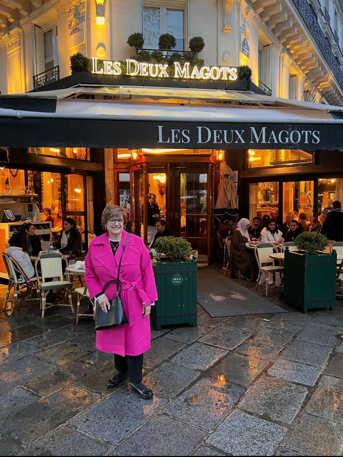 A woman in a bright pink coat outside Les Deux Magots café in Paris at dusk